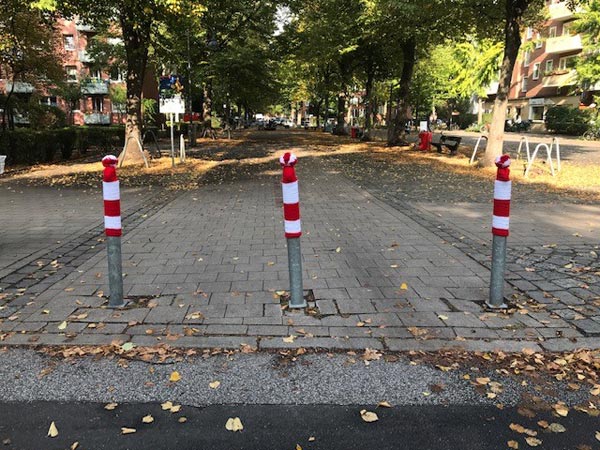 3 graue Poller mit rot-weiß-gestreiften Mützen vor einem Fussgängerbereich mit grauen Steinen