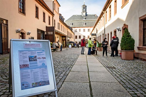 Rathaus Burgstädt - links und rechts am Bildrand befinden sich lange Häuser, welche zum Rathaus im Hintergrund führen. Auf dem gepflasterten Platz zwischen den Häusern sind auf der rechten Seite Biertischgarnituren aufgebaut, an denen Personen sitzen. Auf der rechten Seite stehen blinde und sehebehinderte Menschen und unterhalten sich mit dem Security-Personal. Außerdem stehen dort große Pflanzenkübel mit Buchsbäumen. Am untern linken Bildrand steht ein Aufsteller mit den Hinweisen zu den beiden Aktionstagen.