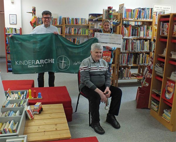 Bibliothek Burgstädt - Übergabe der Geldspende an die Leiterin der Kinderarche - Burgstädt. In der Mitte des Bildes sitzt Olaf Schmidt auf einem Stuhl. Dahinter steht der Bürdermeister von Burgstädt und die Leiterin der Kinderarche. Beide halten gemeinsam das grünweiße Banner der Kinderache in der Hand. Die Leiterin hält auußerdem den Spendenscheck in ihrer linken Hand. Im Hintergrund und auf der rechten Seite stehen gefüllte Bücherregale. Auf der linken Seite stehen vier Bücherkisten, eine Holzkiste und drei Stapel mit roten quadratischen Matratzen.