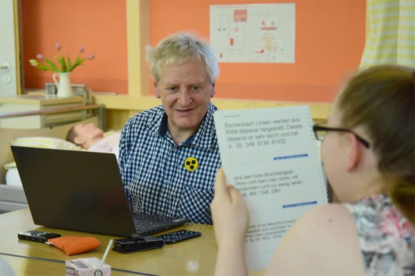 In einem Lehrzimmer der Carus Akademie, sitzen ein blinder Mann und eine Auszubildende gemeinsaman einem Tisch. Vor dem Mann steht ein Laptop und die Schülerin lässt sich einen Text mit der Orcam vorlesen. Im Hintergrund steht ein Pflegebett mit einem Dummy.