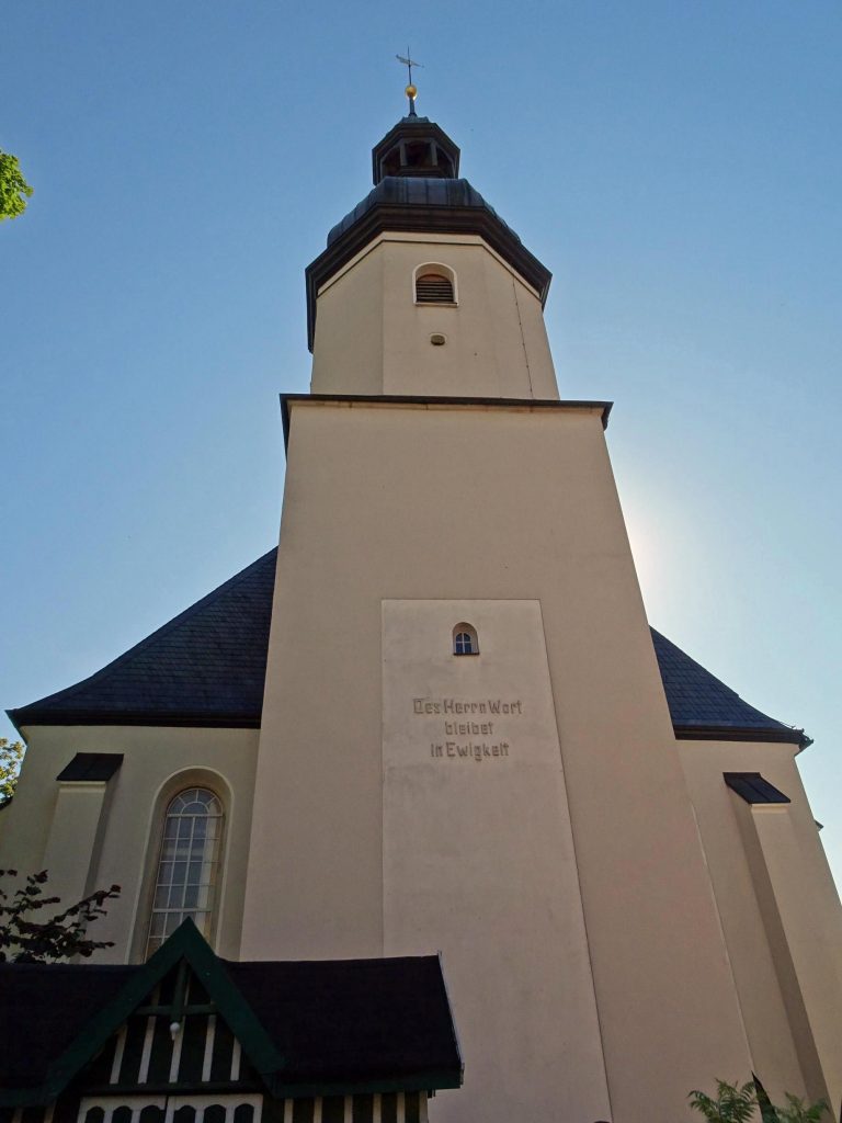 Kirche mit dem Schriftzug "Des Herrn Wort bleibt in Ewigkeit". Im Hintergrund blauer Himmel und Sonnenschein.