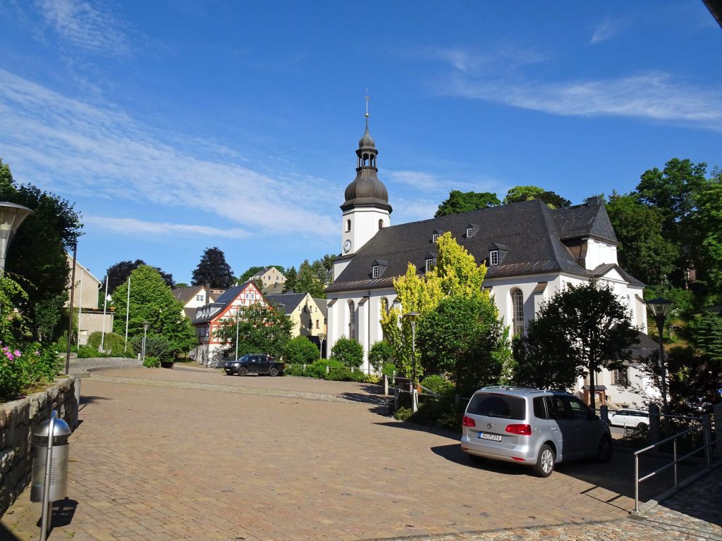 Kirche mit gepflastertem Vorplatz, links und rechts davon stehen Bäume sowie Fachwerkhäuser.