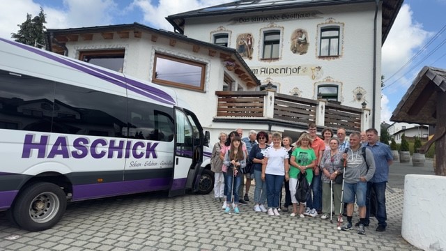 Auf der rechten Seite im Bild stehen die Teilnehmer, links der Kleinreisebus mit dem Schriftzug "Haschick" in Lila. Dahinter ist der das "Klingende Gasthaus Alpenhof". Der Himmel im Hintergrund ist blau, mit großen weißen Wolken.