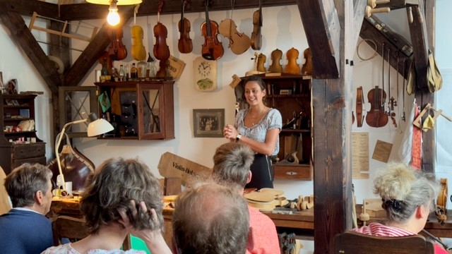 Eine Frau erklärt dem Publikum die einzelnen Arbeitsschritte beim Bau einer Geige. Auf der Werkbank stehen eine weiße Schreibtischlampe und Einzelteile einer Geige. Im Hintergrund an der weißen Wand hängen zwei braune Regale, eine Uhr und Geigen in verschiedenen Fertigungsstufen.

