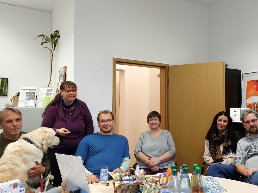 Von links nach rechts auf dem Bild zu sehen: Mike Jakob mit seinem Assistenzhund Ben, einem Golden Retriever, auf dem Schoß. Daneben Jana Färber stehend mit dem Kamera-Auslöser in der Hand. Dann Martin Siebenstädt, Beate Schilbach, Katja Oltersdorf und Daniel Martin. Andreas Müller ist nicht mit auf dem Foto. Die Teilnehmer sitzen um einen Tisch herum, auf dem Getränke, Snacks und einige Büro-Utensilien stehen. Links in der Ecke des Raums steht eine hohe Grünpflanze.  Links daneben ist ein Teil eines grauen Schranks zu sehen, auf dem Infomaterial angeordnet ist. An den Wänden hängen Bilder und Plakate. Die Tür zum Flur ist offen. 