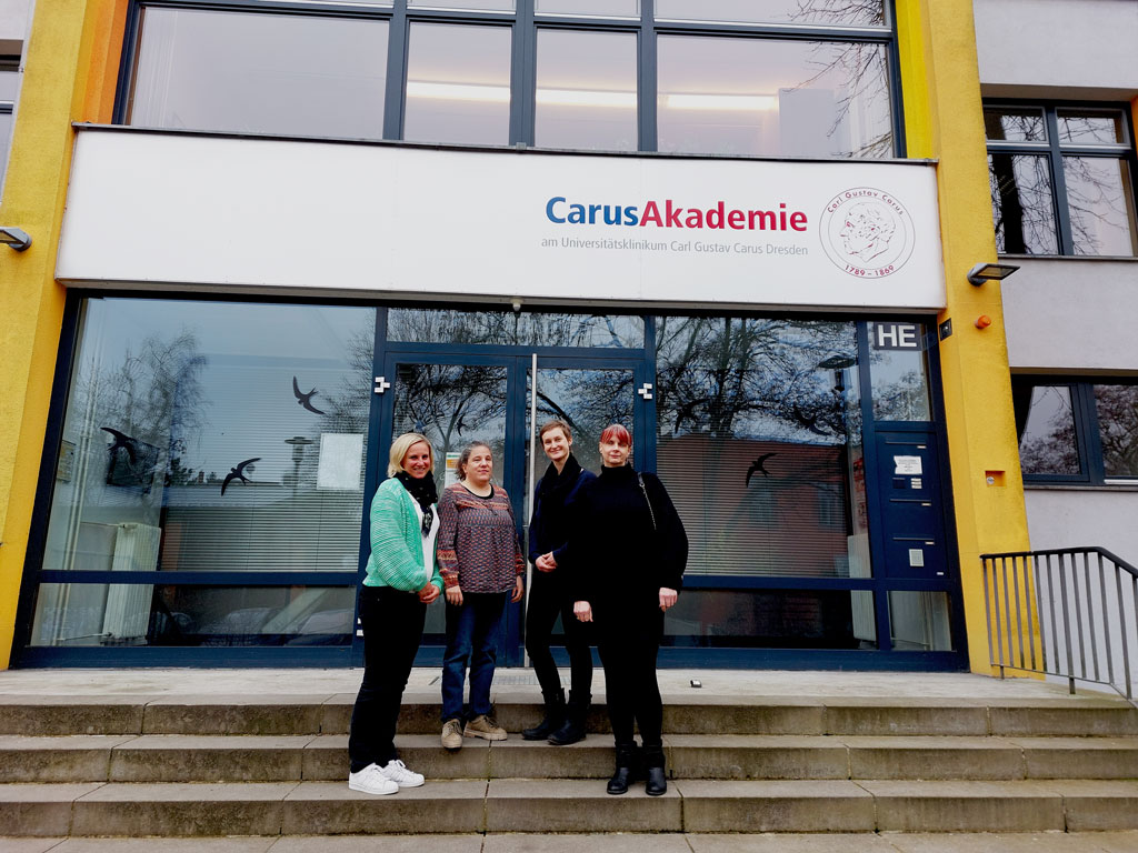 Vor dem Eingang zur CarusAkademie stehen auf der Treppe zwei Beraterinnen von Blickpunkt Auge, eine Hilfsmittelberaterin aus dem Landeshilfsmittelzentrum und eine Assistenzkraft.