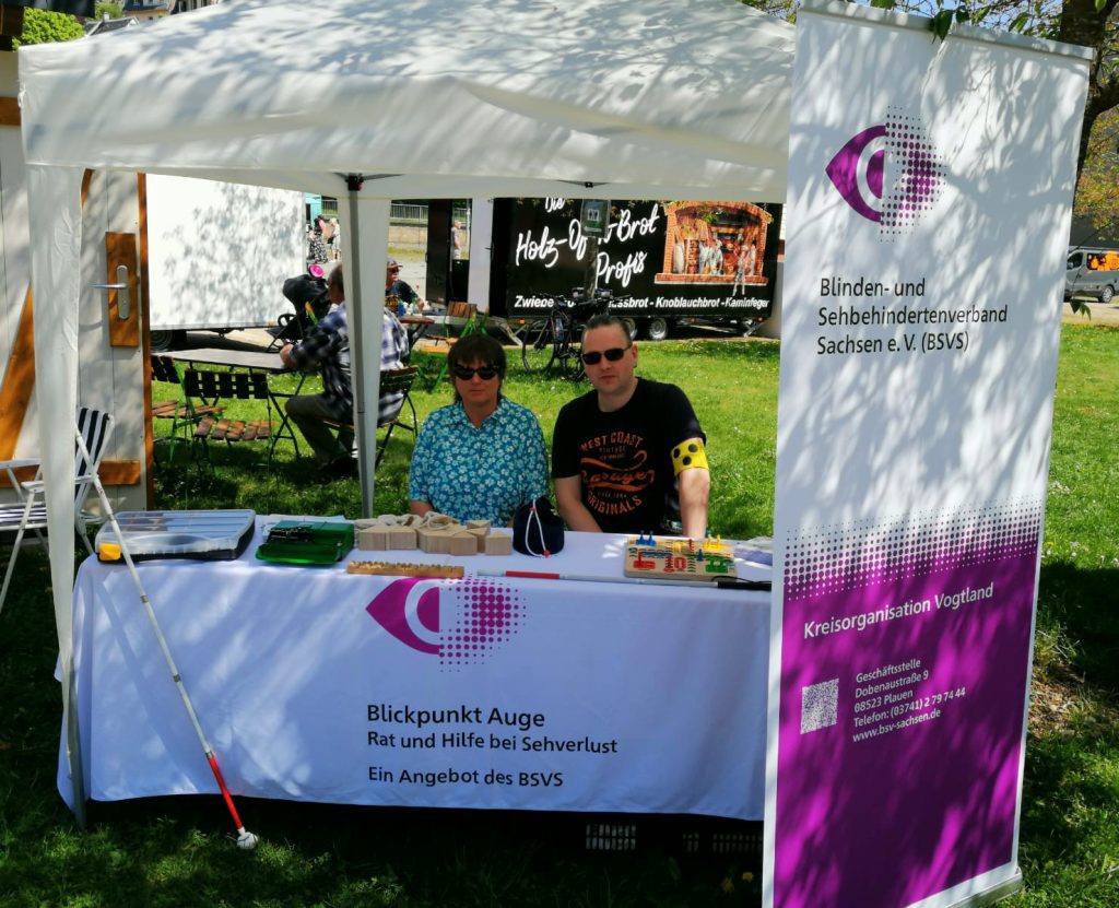 Frau Beate Schilbach und Herr Daniel Martin sitzen am Stand der Kreisorganisation Vogtland unter einem Pavillon auf einer Wiese. Auf der rechten Seite steht das Roll-UP der KO Vogtland und links ist ein Langstock am Stand angelehnt. Im Hintergrund stehen andere Informationsstände.