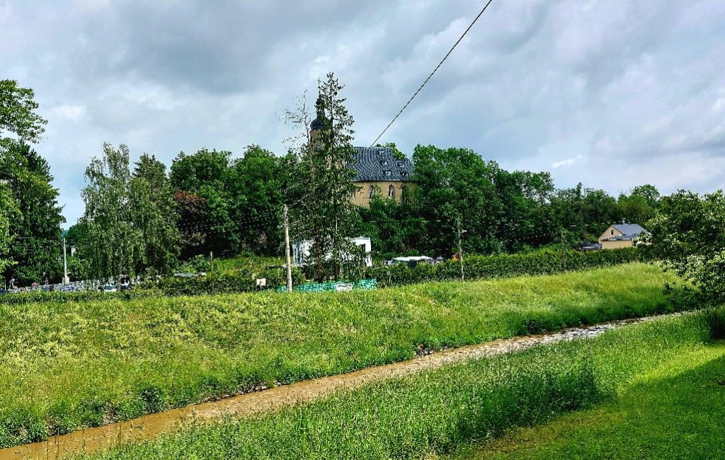 Blick auf die Straßberger Kirche. Im Vordergrund fließt die Weiße Elster. Das Wasser ist trüb und schlammig.
