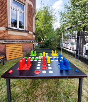 Das Foto zeigt einen großen Holztisch, in dem das Mensch-Ärgere-Dich-Nicht-Spiel eingearbeitet ist, welches neben unserem Gebäude im grünen Beratungsraum steht. Die Spielsteine und Würfel sind sehr groß, kontrastreich und taktil gestaltet.