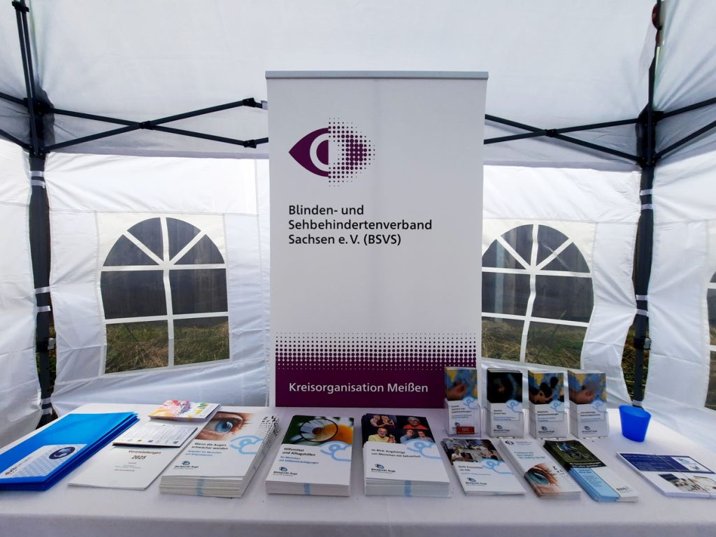 Das Foto zeigt einen Infostand des Blinden- und Sehbehindertenverbandes Sachsen e. V. (BSVS), Kreisorganisation Meißen. Hinter dem Tisch steht ein Roll-up mit Logo und Verbandsname. Auf dem Tisch liegen geordnet verschiedene Flyer, Broschüren und Informationsmaterialien, daneben ein blauer Becher. Der Stand befindet sich in einem weißen Pavillon.