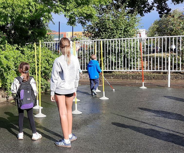 Das Bild zeigt drei Kinder im Freien auf einem nassen Asphaltplatz. Ein Junge läuft mit einem weißen Langstock durch einen Parcours aus bunten Stangen. Zwei Mädchen stehen daneben und beobachten ihn, eines trägt einen Rucksack, das andere kurze Hose und grauen Pullover. Im Hintergrund sind ein weißer Zaun, Büsche und Gebäude zu sehen.