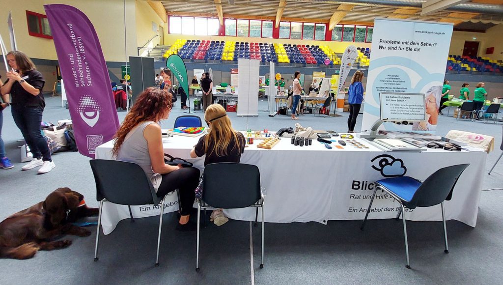 Das Bild zeigt einen Stand von Blickpunkt Auge in einer Sporthalle. Im Vordergrund sitzt eine Frau mit einem Kind, das eine Augenbinde trägt, an einem Tisch mit verschiedenen Spielen und Hilfsmitteln für Sehbehinderte. Ein Blindenhund liegt ruhig am Boden neben dem Tisch. Der Stand ist informativ gestaltet und bietet Beratung zu Problemen mit dem Sehen. Im Hintergrund sind weitere Stände und Besucher zu sehen. Das Gesamtbild vermittelt einen Eindruck von Hilfsbereitschaft und Unterstützung für Menschen mit Sehbehinderungen.