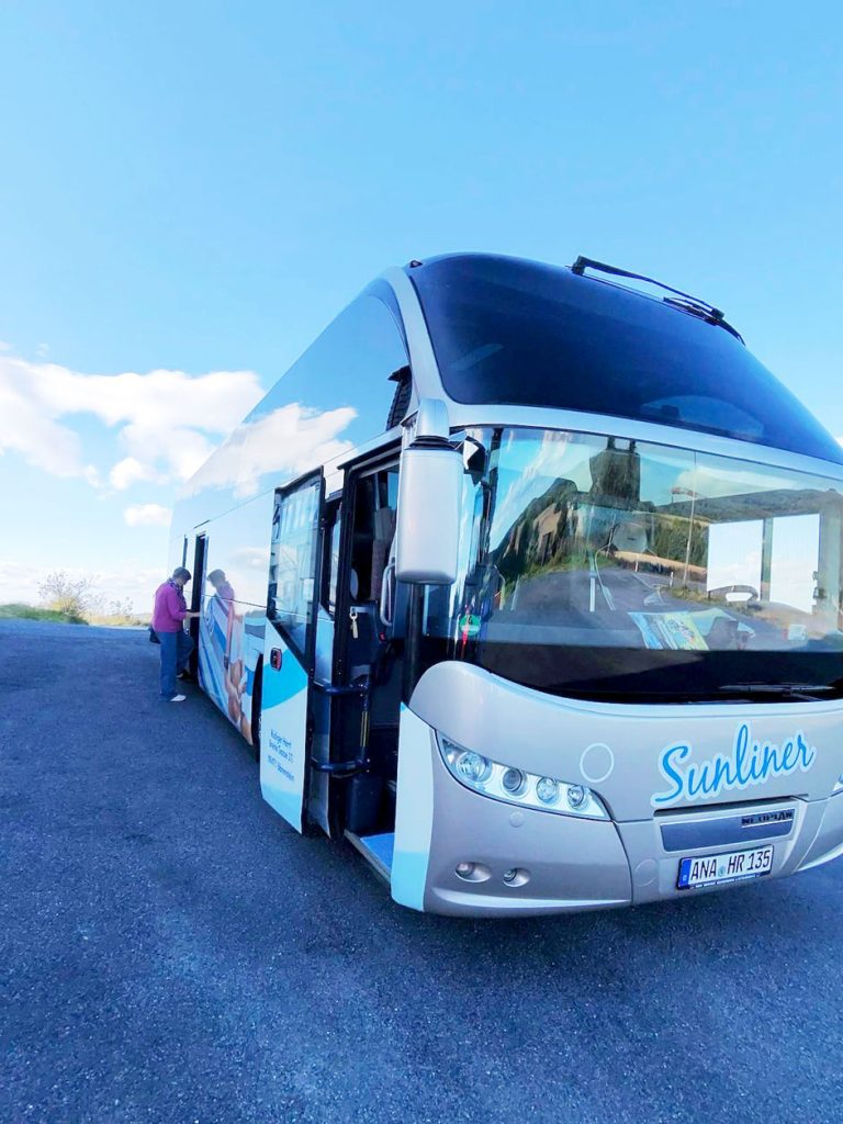 Ein moderner, silberner Reisebus mit der Aufschrift „Sunliner“ steht auf einem Parkplatz unter blauem Himmel. Eine Person mit pinkfarbener Pullover steigt in den Bus.