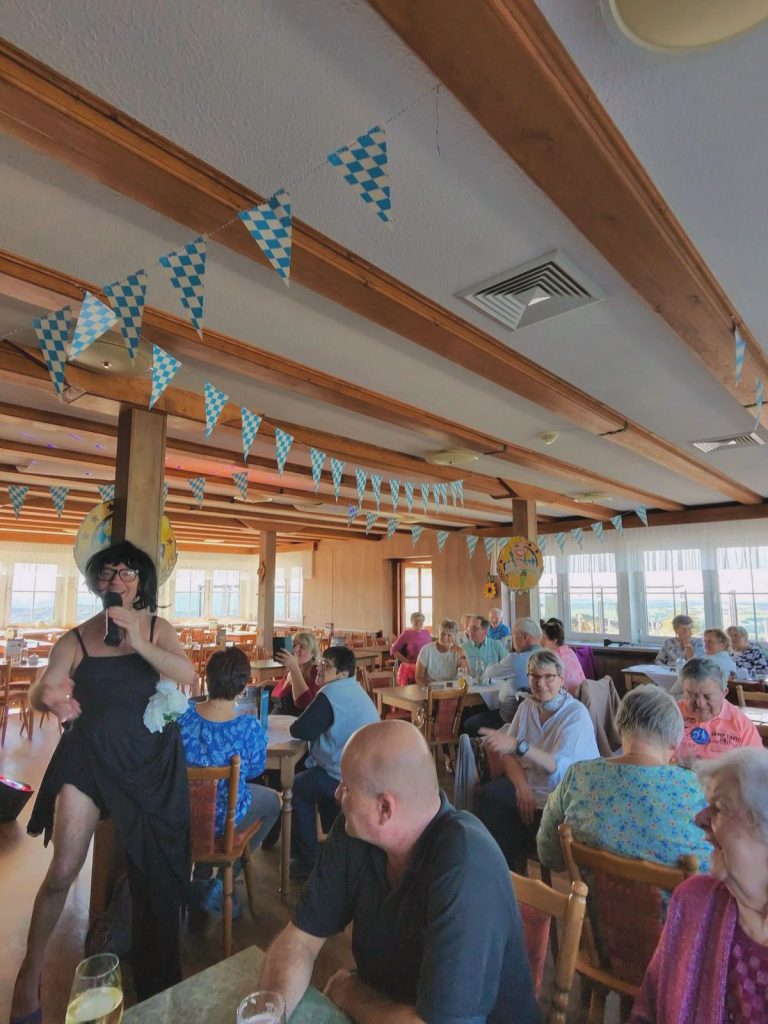 In einem festlich geschmückten Saal mit blau-weißen Wimpelgirlanden sitzen viele Menschen an Tischen und lachen. Eine Person in schwarzem Kleid und Perücke sorgt mit einem Mikrofon in der Hand für Unterhaltung. Die Stimmung wirkt ausgelassen und heiter.