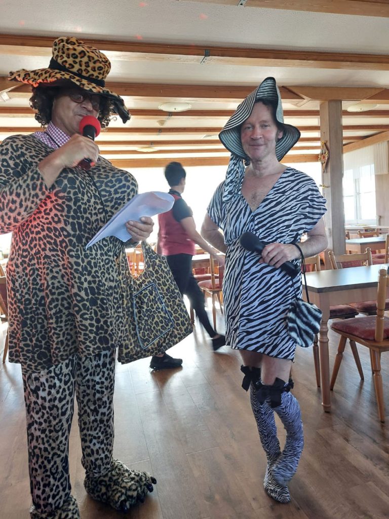 Person in Ganzkörper-Löwenkostüm mit Leopardenmuster und Person in Zebra-Kleid mit passendem Hut, Stulpen und Mikrofon in einem Raum mit Holzmöbeln.