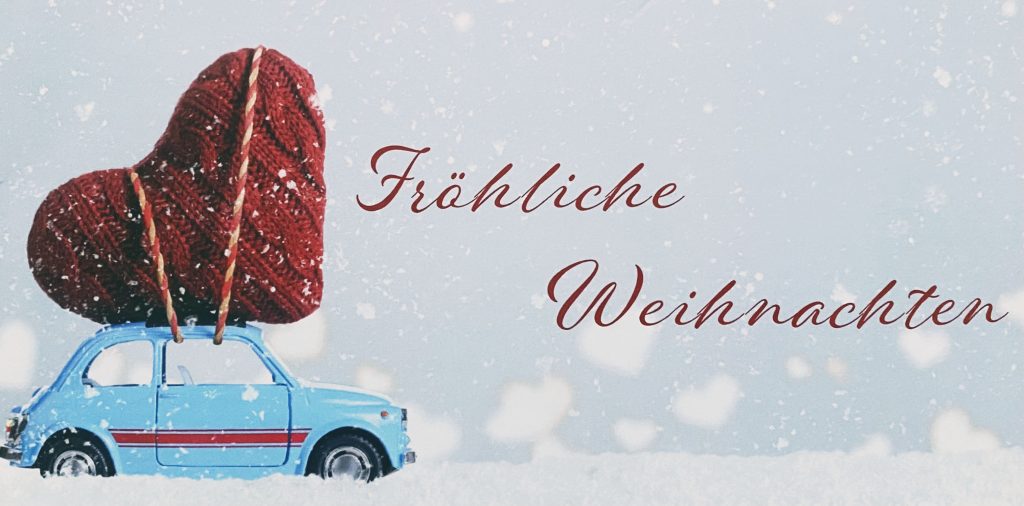 Von links nach rechts fährt ein kleines hellblaues Auto im Schnee, welches auf der Seite einen breiten sowie dünnen roten Streifen hat und auf dem Dach ein festgeschnalltes rotes großes Herz aus Wolle. Es schneit und rechts steht der Schriftzug „Fröhliche Weihnachten“ in Rot.