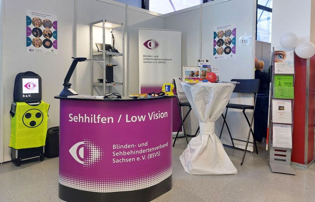 Messestand „Sehhilfen / Low Vision“ des Blinden- und Sehbehindertenverbandes Sachsen e. V. mit Informationsmaterial, vergrößernden Sehhilfen und technischen Hilfsmitteln; im Hintergrund Stellwände mit Plakaten und Sitzgelegenheiten.